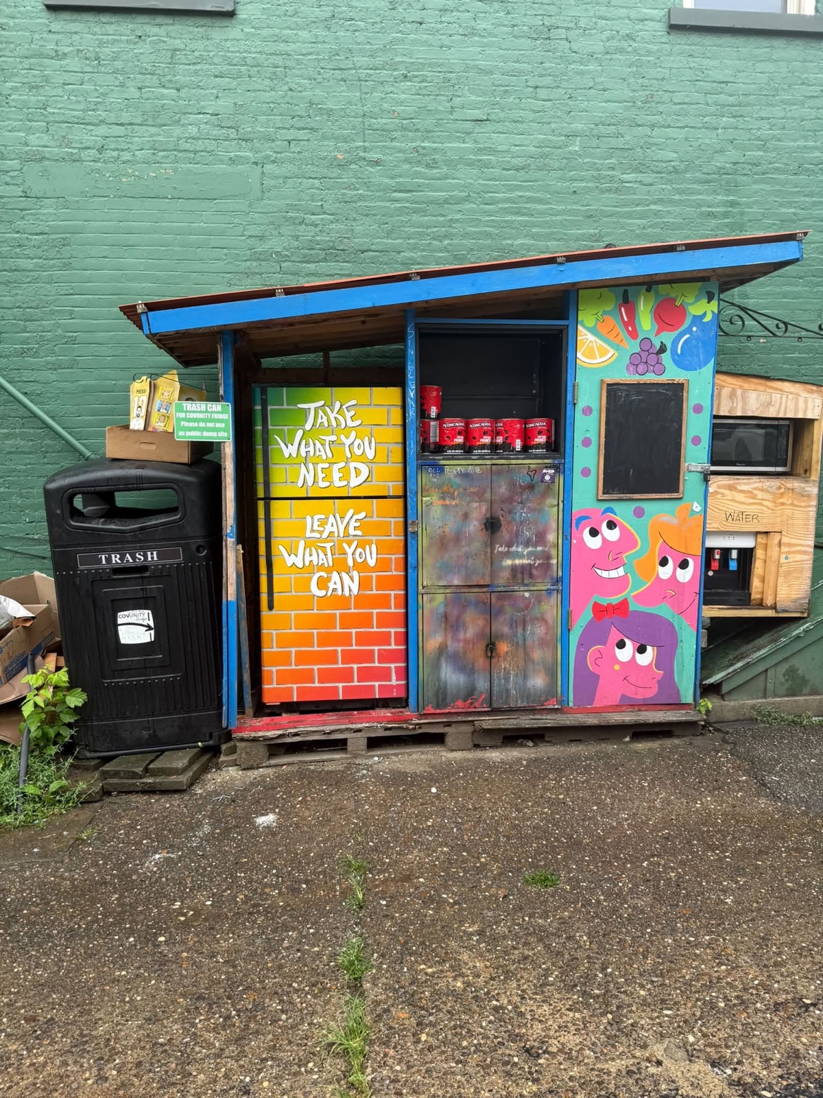The COVUnity Fridge at Redden Gardens: the painted take-what-you-need fridge, pantry cabinet, produce mural panel, and water station under a blue awning.